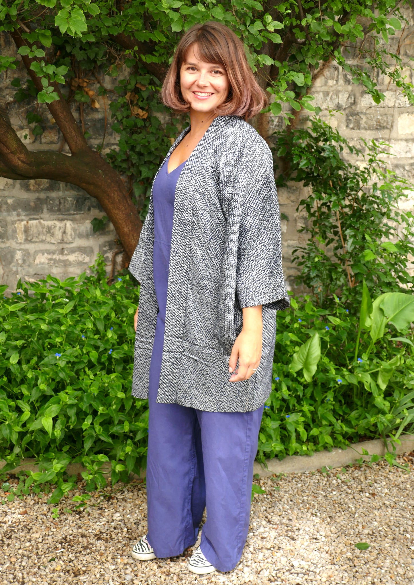 Mikan souriante portant une veste kimono haori bleu-gris à motifs shibori, en soie japonaise, coupe ample et élégante. Look décontracté dans un jardin naturel, avec combinaison bleue et sneakers zébrées.