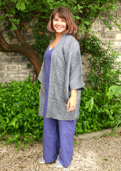 Mikan souriante portant une veste kimono haori bleu-gris à motifs shibori, en soie japonaise, coupe ample et élégante. Look décontracté dans un jardin naturel, avec combinaison bleue et sneakers zébrées.