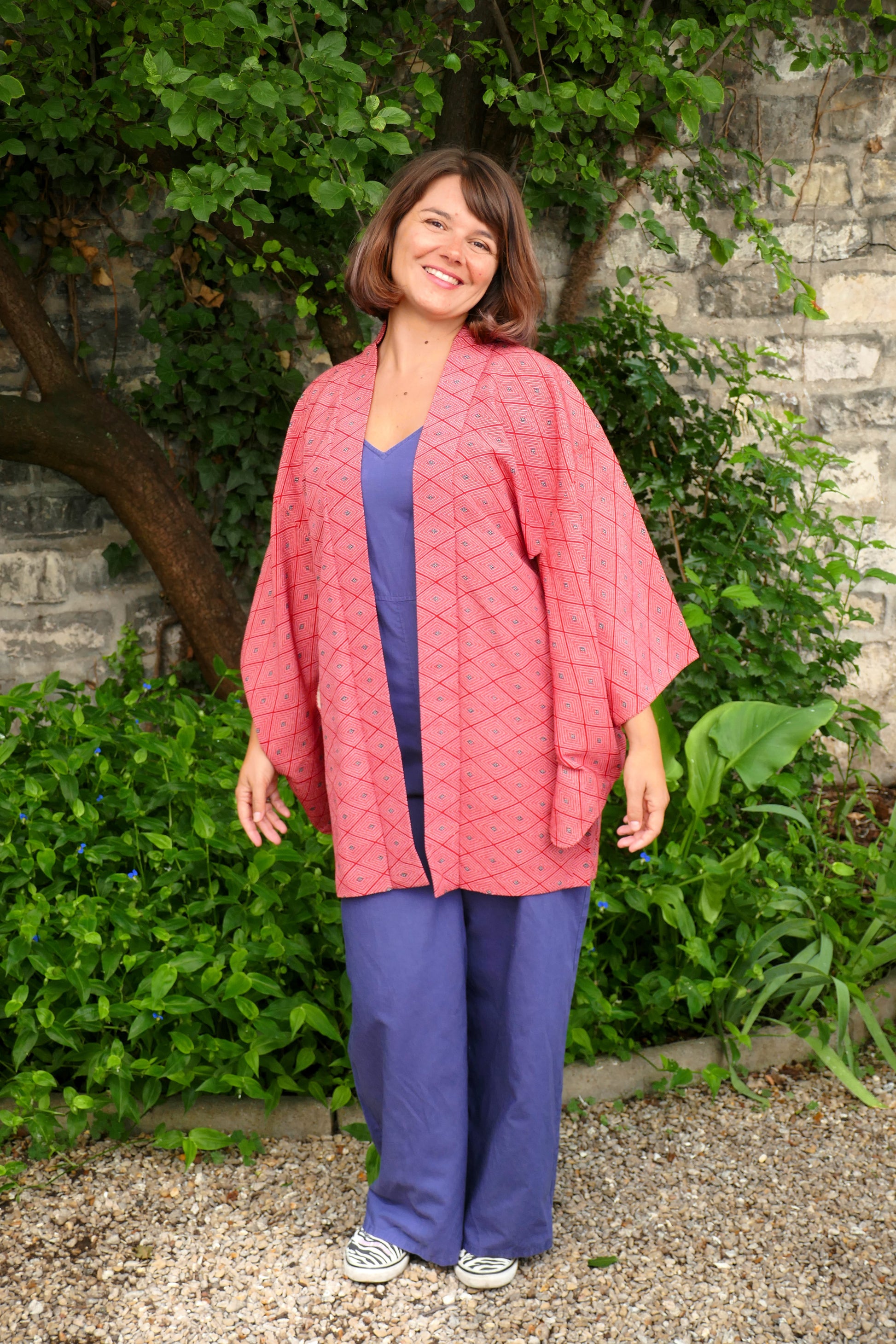 Femme souriante portant une veste de style kimono haori rouge à motifs losange, en extérieur devant un mur de pierre et des plantes vertes.
