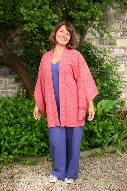 Femme souriante portant une veste de style kimono haori rouge à motifs losange, en extérieur devant un mur de pierre et des plantes vertes.
