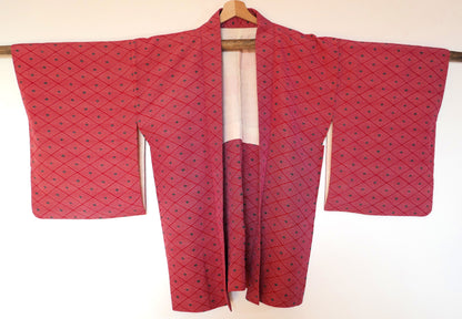 Haori vintage en soie rouge suspendu sur un cintre, montrant le motif géométrique en losanges et la coupe traditionnelle.