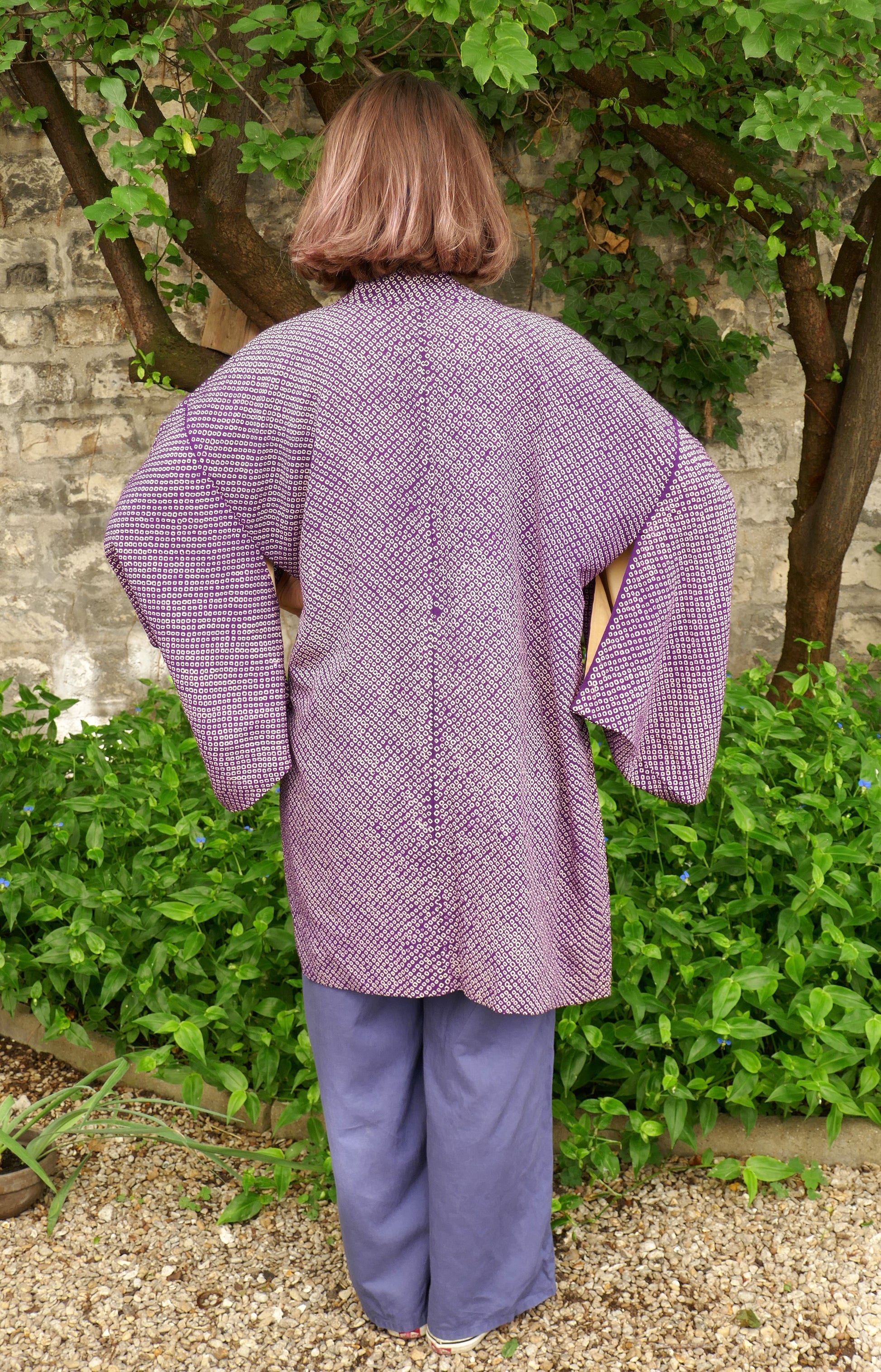 Mikan de dos portant un haori violet vintage à motif shibori, debout dans un jardin fleuri, devant un mur en pierre naturelle. Veste kimono ample en soie japonaise traditionnelle.