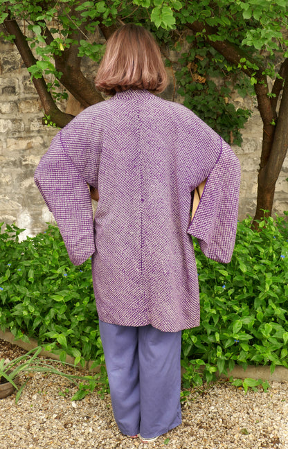 Mikan de dos portant un haori violet vintage à motif shibori, debout dans un jardin fleuri, devant un mur en pierre naturelle. Veste kimono ample en soie japonaise traditionnelle.