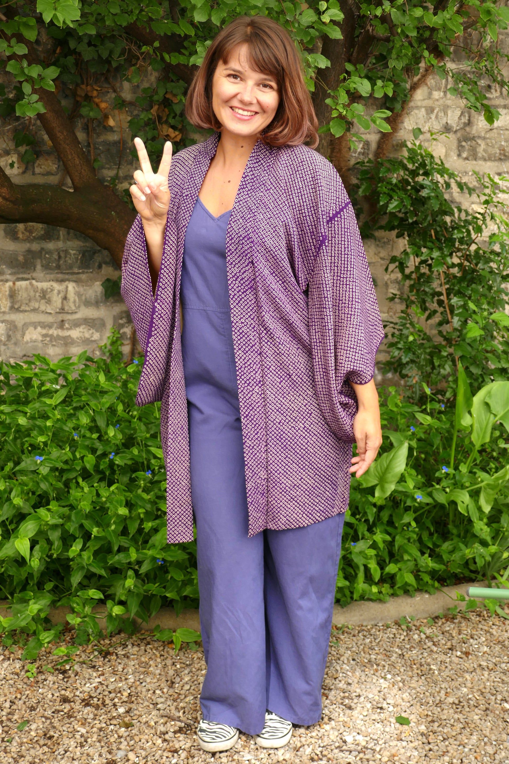 Mikan souriante en haori japonais violet à imprimé shibori, signe de paix avec les doigts, tenue décontractée dans un jardin verdoyant. Veste kimono vintage pour look bohème chic.