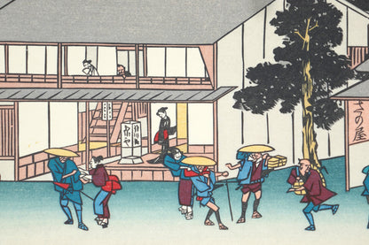 Scène animée devant une auberge à Minakuchi : voyageurs, porteurs et passants dans un décor urbain d’époque Edo.