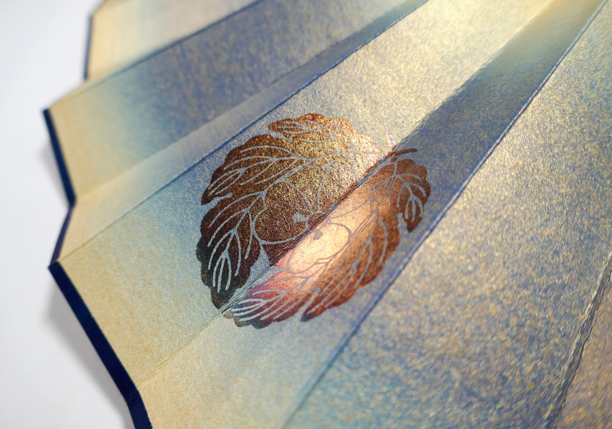 Détail du kamon doré sur papier irisé bleu et or d’un éventail japonais décoratif de collection.