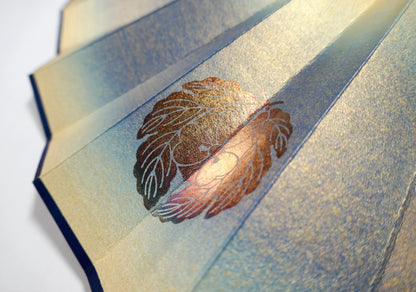 Détail du kamon doré sur papier irisé bleu et or d’un éventail japonais décoratif de collection.