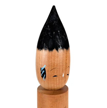 Gros plan du visage : kokeshi fude en bois, expression sereine, détails peints (yeux, sourcils, mèches bleutées), objet japonais vintage.