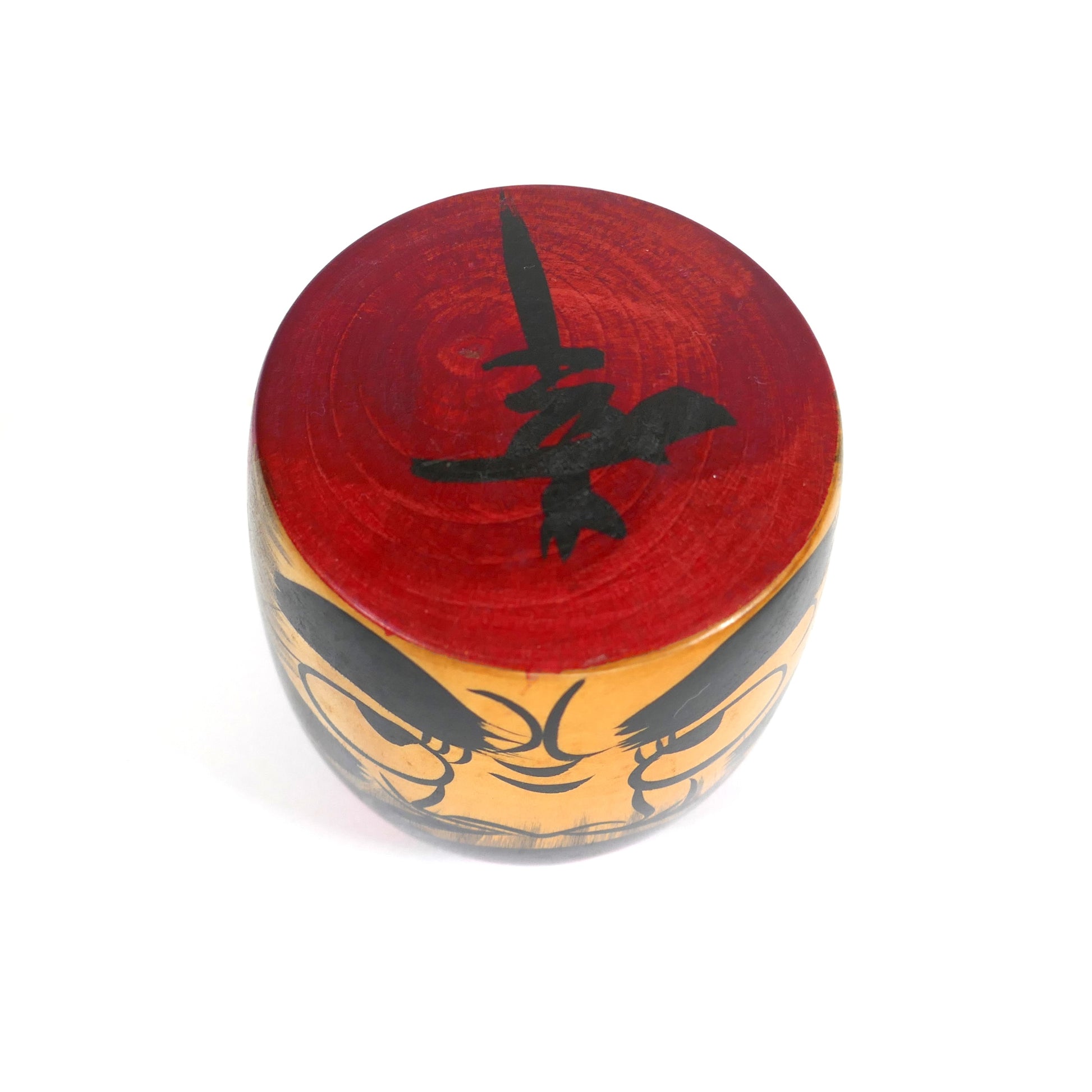 Vue du dessus d’une kokeshi Daruma, avec kanji noir sur fond rouge peint
