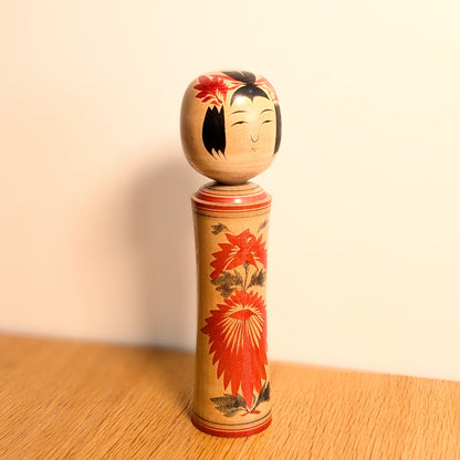 Kokeshi japonaise traditionnelle Naruko d’Okazaki Seishi présentée en décoration, avec motif hishigiku rouge sur bois naturel.