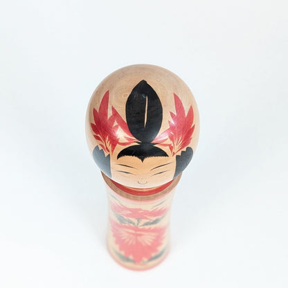 Vue du dessus d’une kokeshi Okazaki Seishi montrant la coiffure peinte et les ornements rouges d’une poupée traditionnelle Naruko.