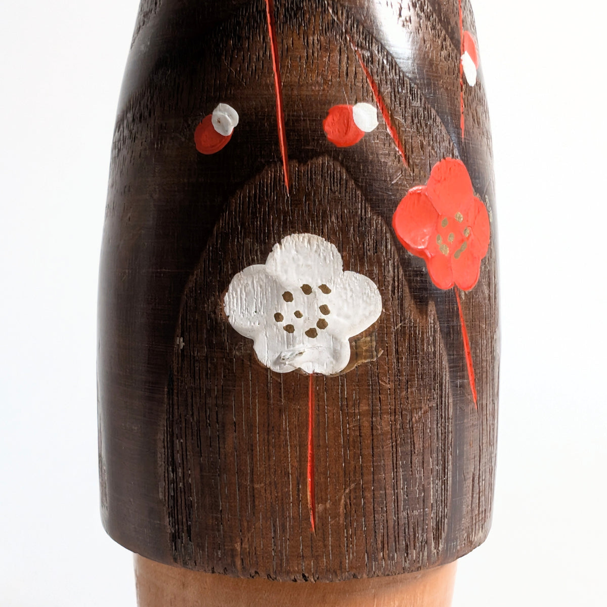 Gros plan des fleurs de prunier ume rouges et blanches peintes en relief sur le corps brun foncé d'une kokeshi sōsaku japonaise, avec la texture du bois visible.
