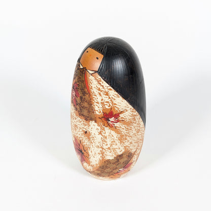 Vue latérale de la kokeshi Sōsaku Usaburo, avec coiffure noire gravée et kimono moucheté orné de feuilles d’érable rouges.