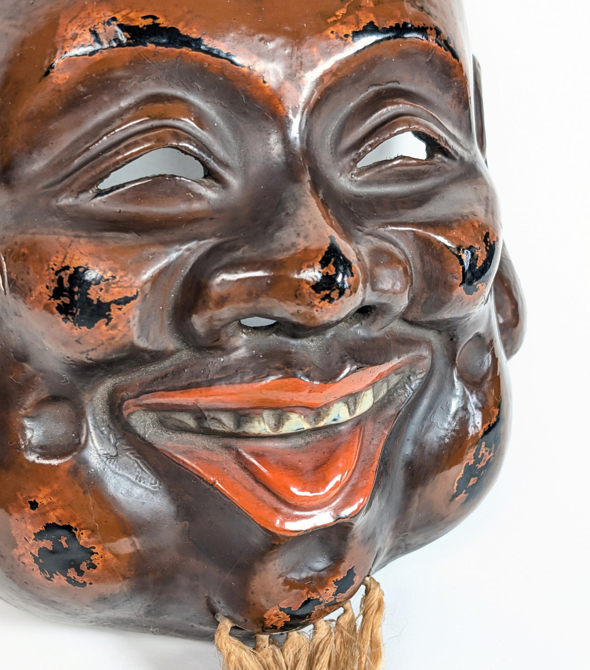 Gros plan sur le sourire d’un masque de Daikoku, avec lèvres et langue vermillon, dents peintes et laque brun-noir patinée.