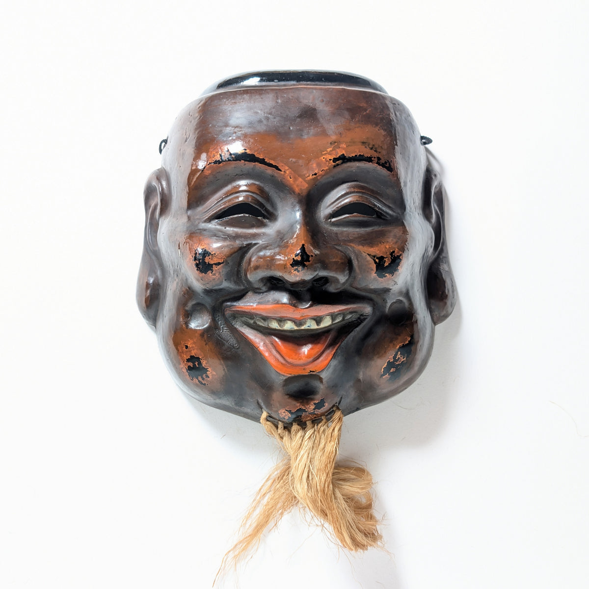 Vue frontale d’un masque japonais de Daikoku, objet d’art traditionnel en laque sèche avec visage souriant et barbe en fibres naturelles.