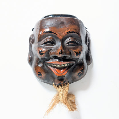 Vue frontale d’un masque japonais de Daikoku, objet d’art traditionnel en laque sèche avec visage souriant et barbe en fibres naturelles.