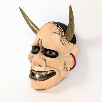 Vue oblique d'un masque Hannya de l'art japonais, avec deux cornes dorées et un large sourire aux crocs pointus.
