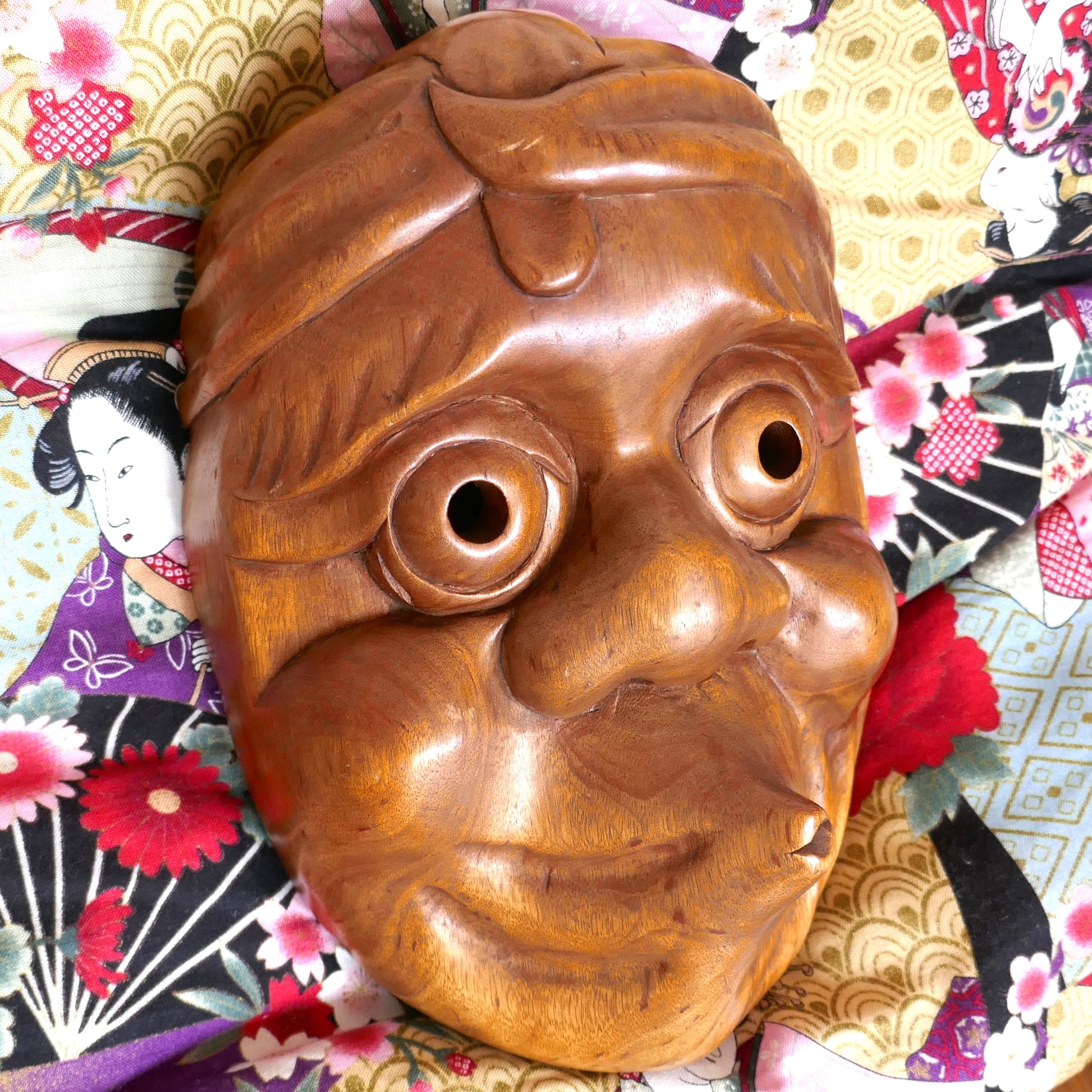 Masque Hyottoko vu en angle gauche, montrant les détails du bois sculpté avec un fond de tissus japonais aux motifs floraux.