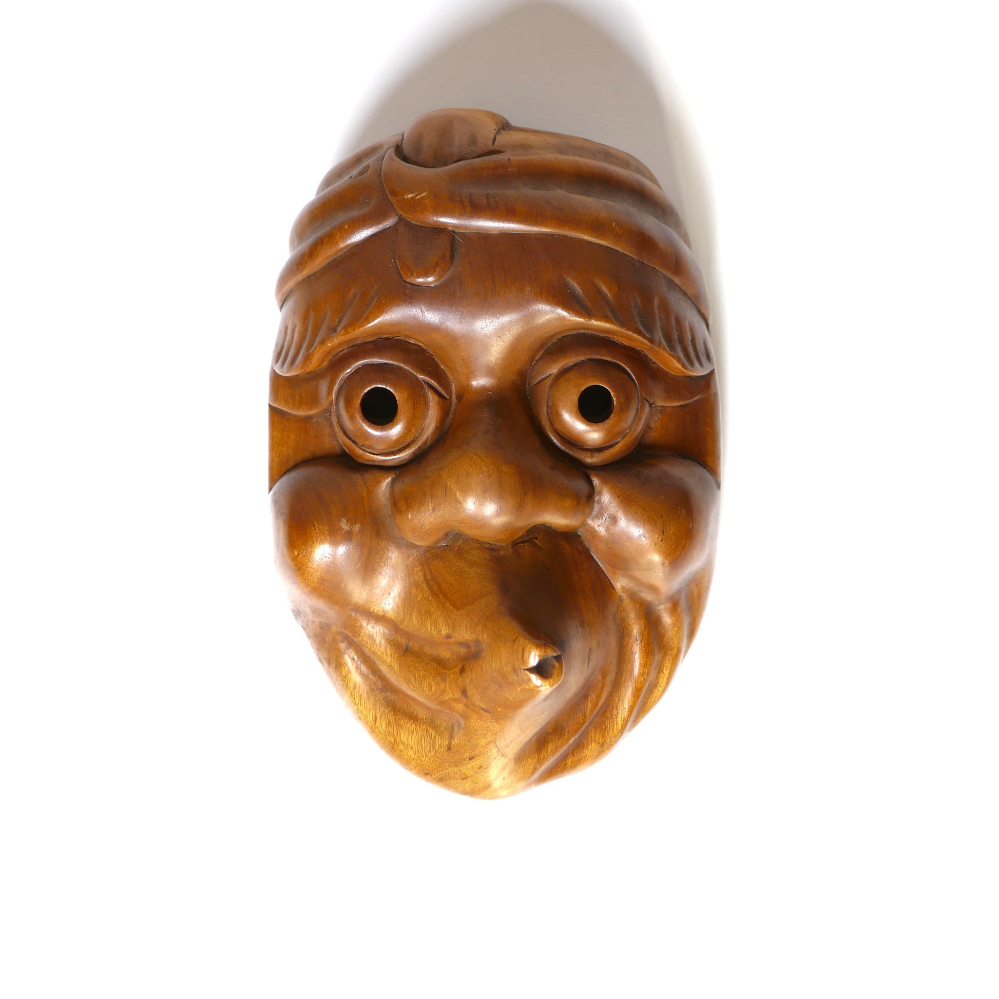 Masque Hyottoko sur fond blanc avec ombre portée, lumière venant du bas, soulignant la texture du bois.