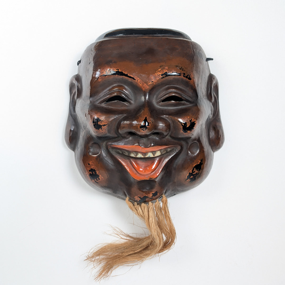 Vue de face d’un masque japonais de Daikoku, une divinité du bonheur, en kanshitsu, laque sèche brun-rouge et noire avec barbe en fibres naturelles.