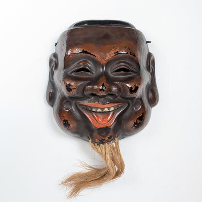 Vue de face d’un masque japonais de Daikoku, une divinité du bonheur, en kanshitsu, laque sèche brun-rouge et noire avec barbe en fibres naturelles.