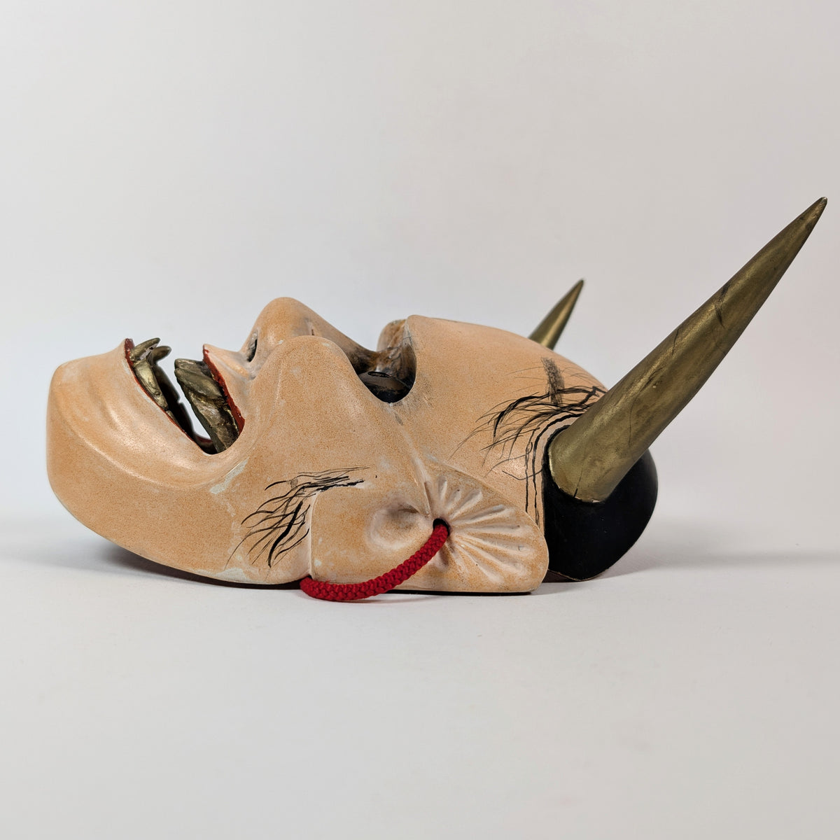 Profil d'un masque traditionnel Nô Hannya posé à plat, avec une corne dorée, l'oreille et le cordon rouge.