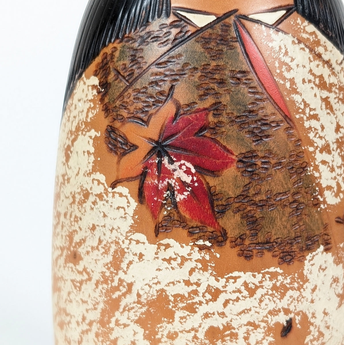 Gros plan d’une feuille de momiji rouge sur le kimono de la kokeshi Usaburo, avec décor brun gravé et peinture blanche mouchetée.