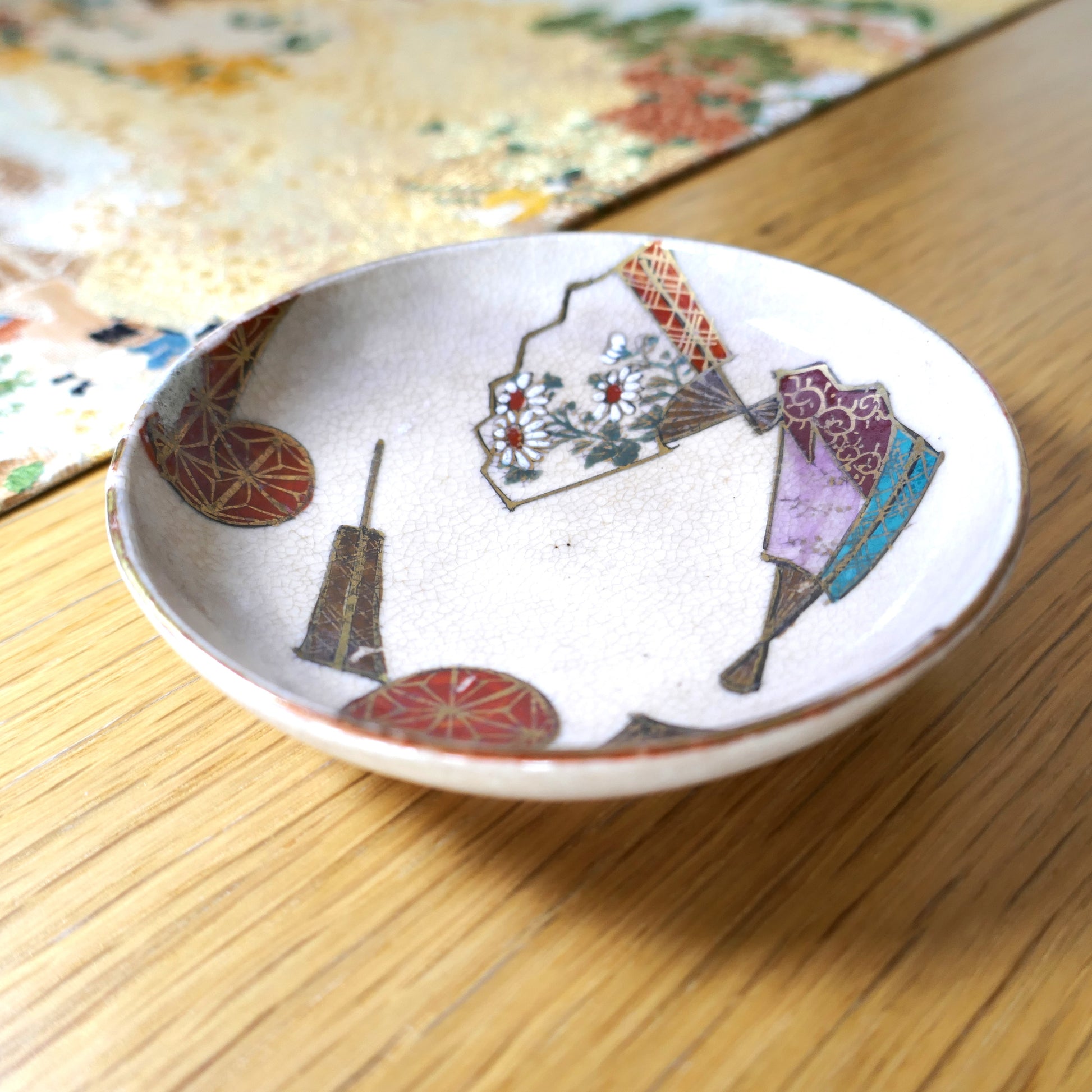Coupelle ancienne japonaise en céramique Kutani, posée sur une table en bois, décorée d’éventails et de fleurs.