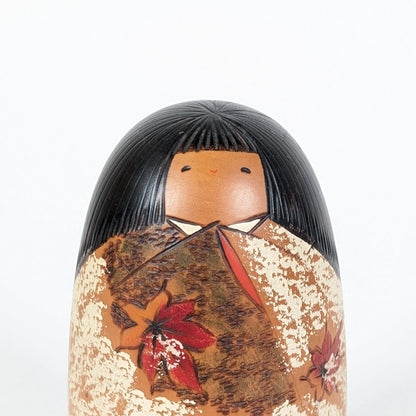 Portrait rapproché de la kokeshi Shôzan : visage doux, frange noire finement sculptée et pans de kimono aux couleurs automnales.