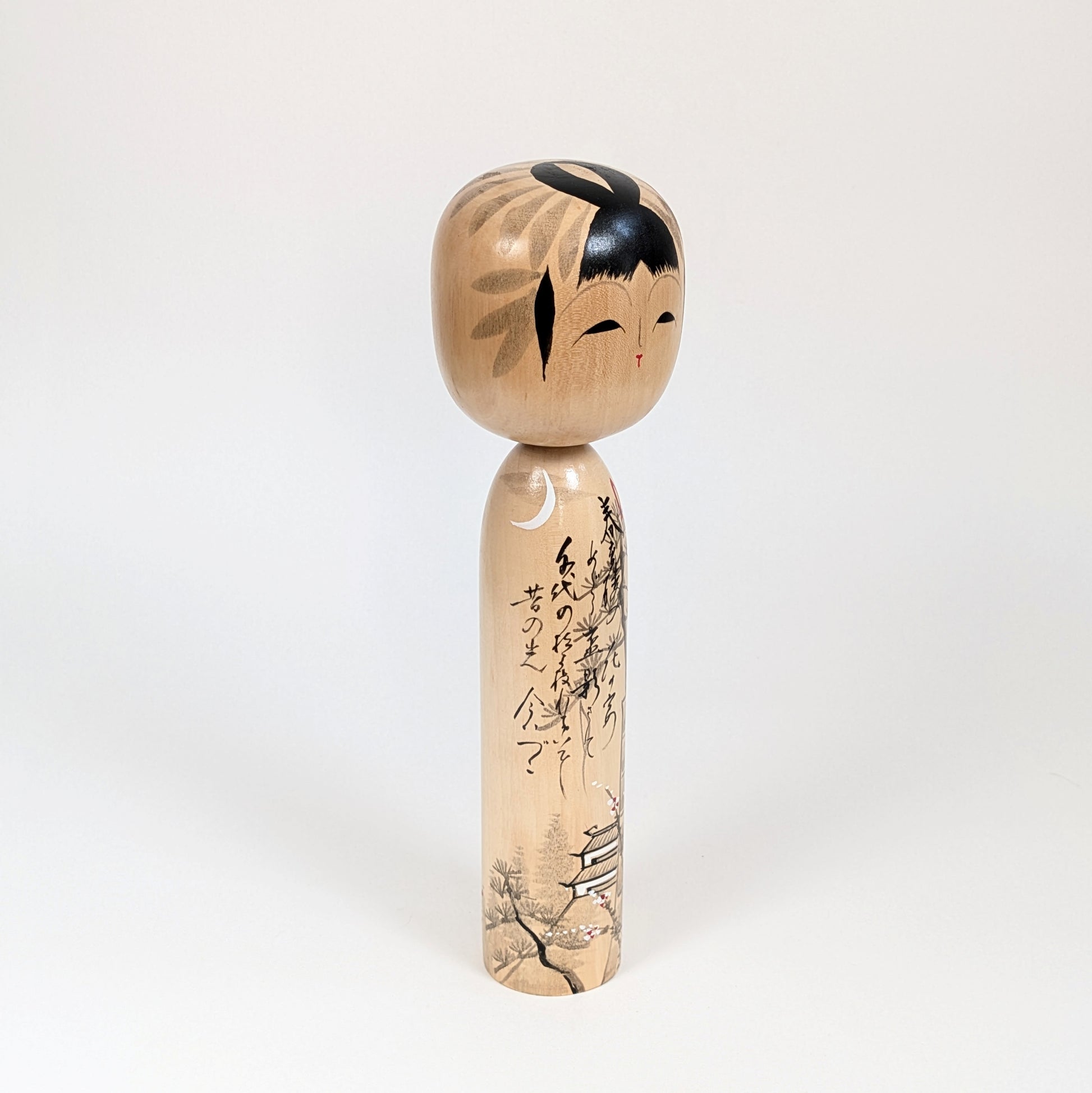 Poupée japonaise en bois signée Shūhō, vue trois quarts avec calligraphie, lune et décor architectural japonais.
