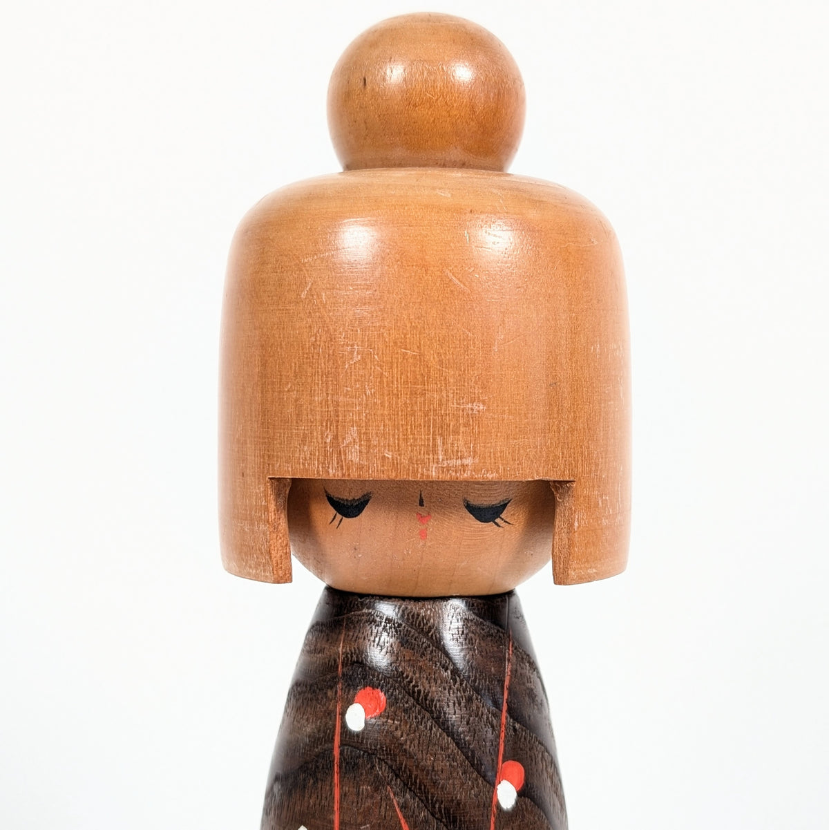 Gros plan du visage délicat de la poupée kokeshi japonaise Koigokoro, avec ses yeux fermés, ses fines paupières peintes et sa coiffure en bois clair surmontée d'un chignon.