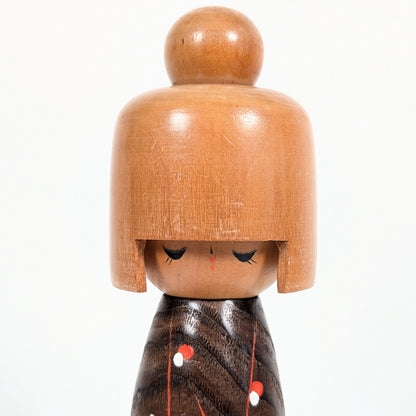 Gros plan du visage délicat de la poupée kokeshi japonaise Koigokoro, avec ses yeux fermés, ses fines paupières peintes et sa coiffure en bois clair surmontée d'un chignon.