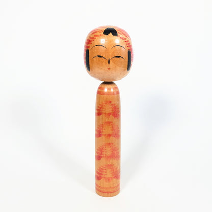 Vue de face d’une kokeshi traditionnelle Togatta signée Onuma Shoji, visage souriant, tête à motif tegara rouge et corps kasanegiku.
