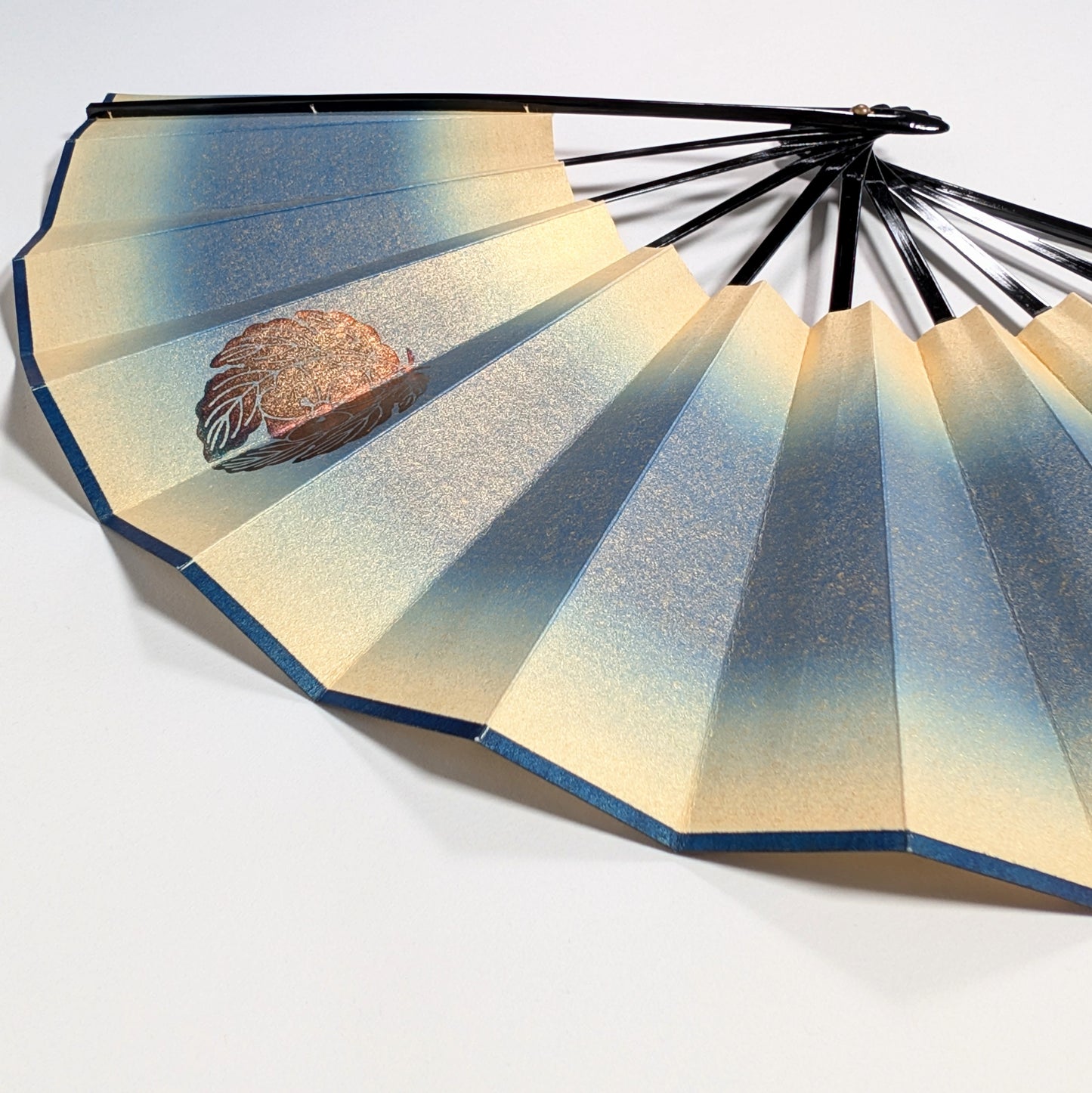 Détail d’un sensu japonais en papier irisé bleu et or avec motif floral doré, éventail pliant traditionnel.
