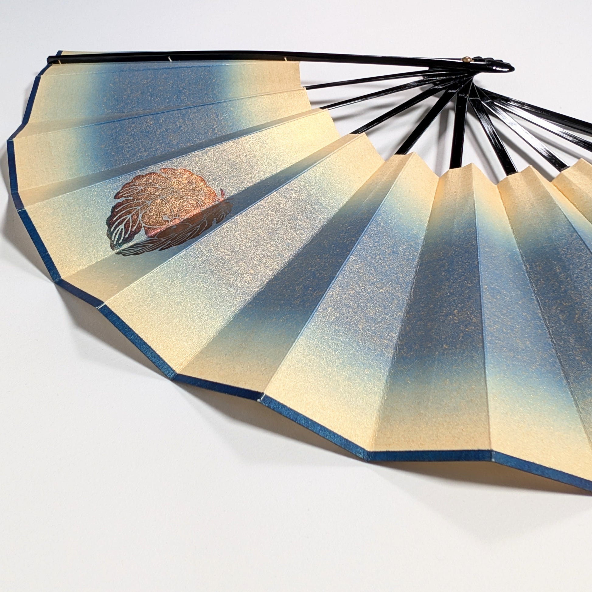 Détail d’un sensu japonais en papier irisé bleu et or avec motif floral doré, éventail pliant traditionnel.