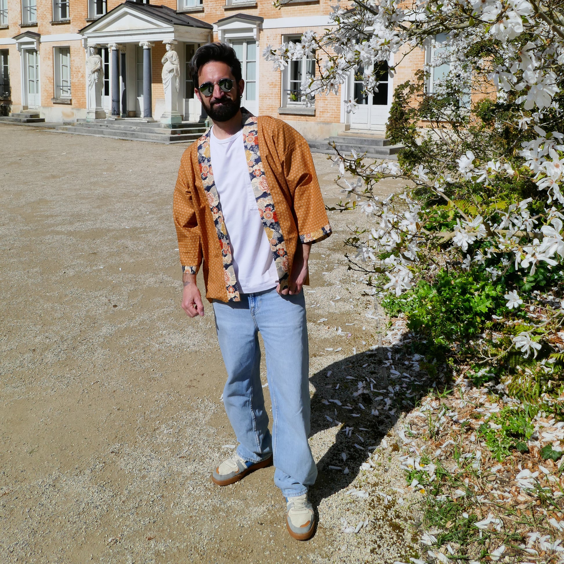 Tôma porte le haori Yūyake Zakura face à l’objectif sous les magnolias en fleurs.


