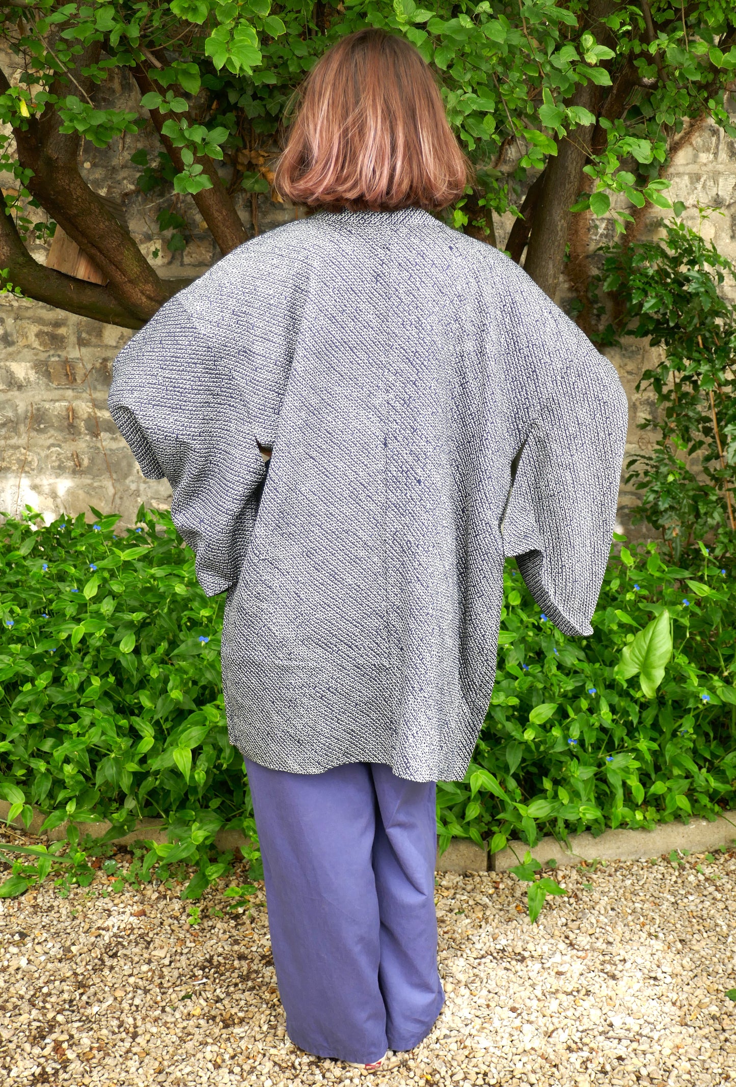 Mikan de dos portant un haori japonais vintage en soie, motif shibori bleu-gris, manches larges type kimono, dans un jardin verdoyant avec mur en pierre. Veste traditionnelle ample et fluide, style wabi-sabi.