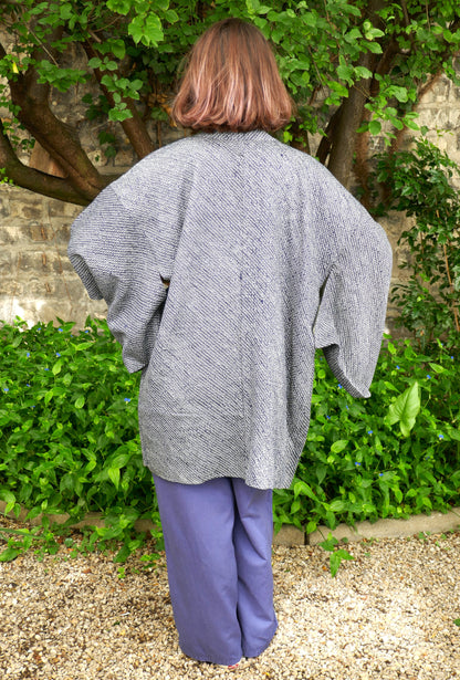 Mikan de dos portant un haori japonais vintage en soie, motif shibori bleu-gris, manches larges type kimono, dans un jardin verdoyant avec mur en pierre. Veste traditionnelle ample et fluide, style wabi-sabi.