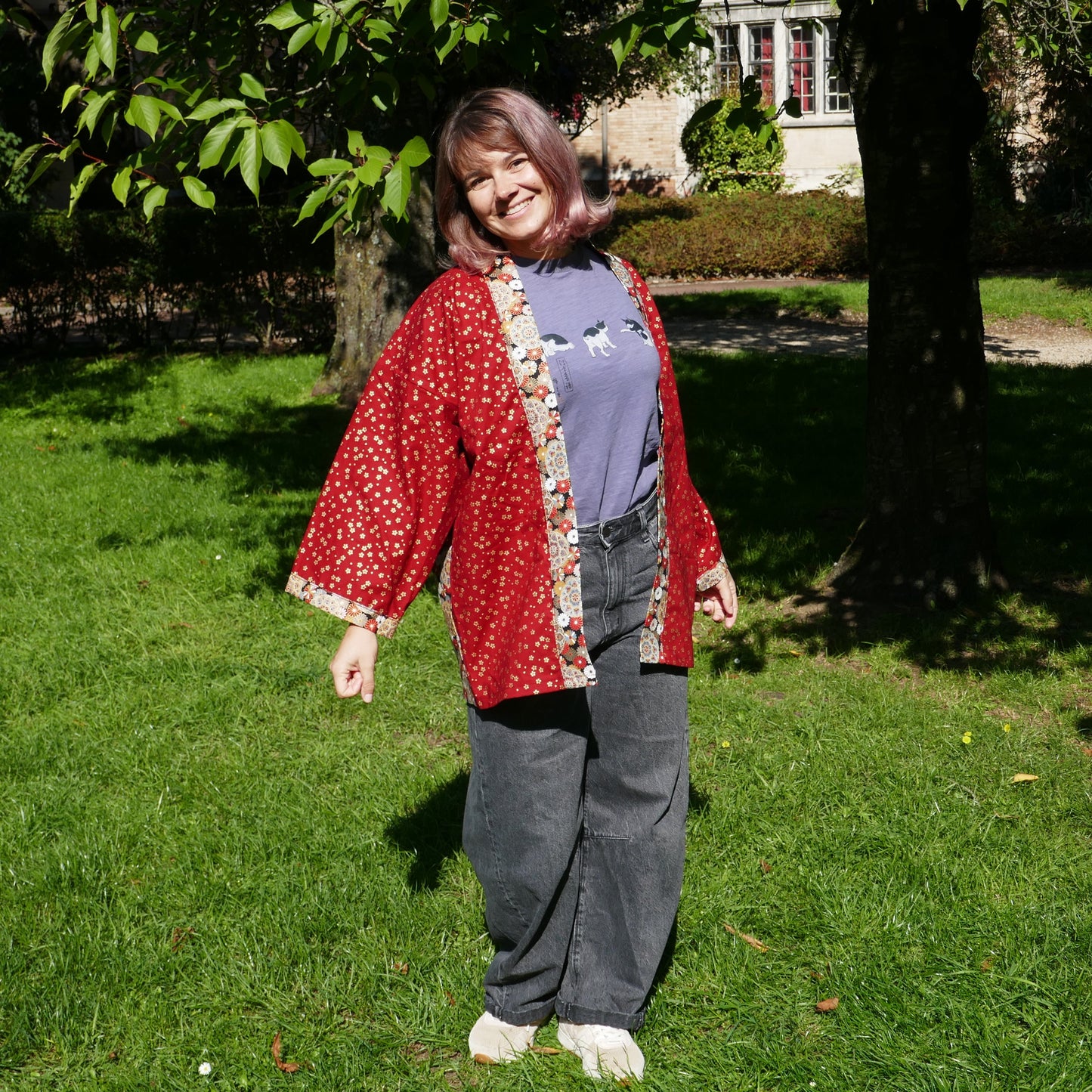 Mikan souriante en veste kimono traditionnelle haori moderne de l'atelier KOGEDO rouge, motif chrysanthèmes 菊 et sakura, dans un parc à Paris.