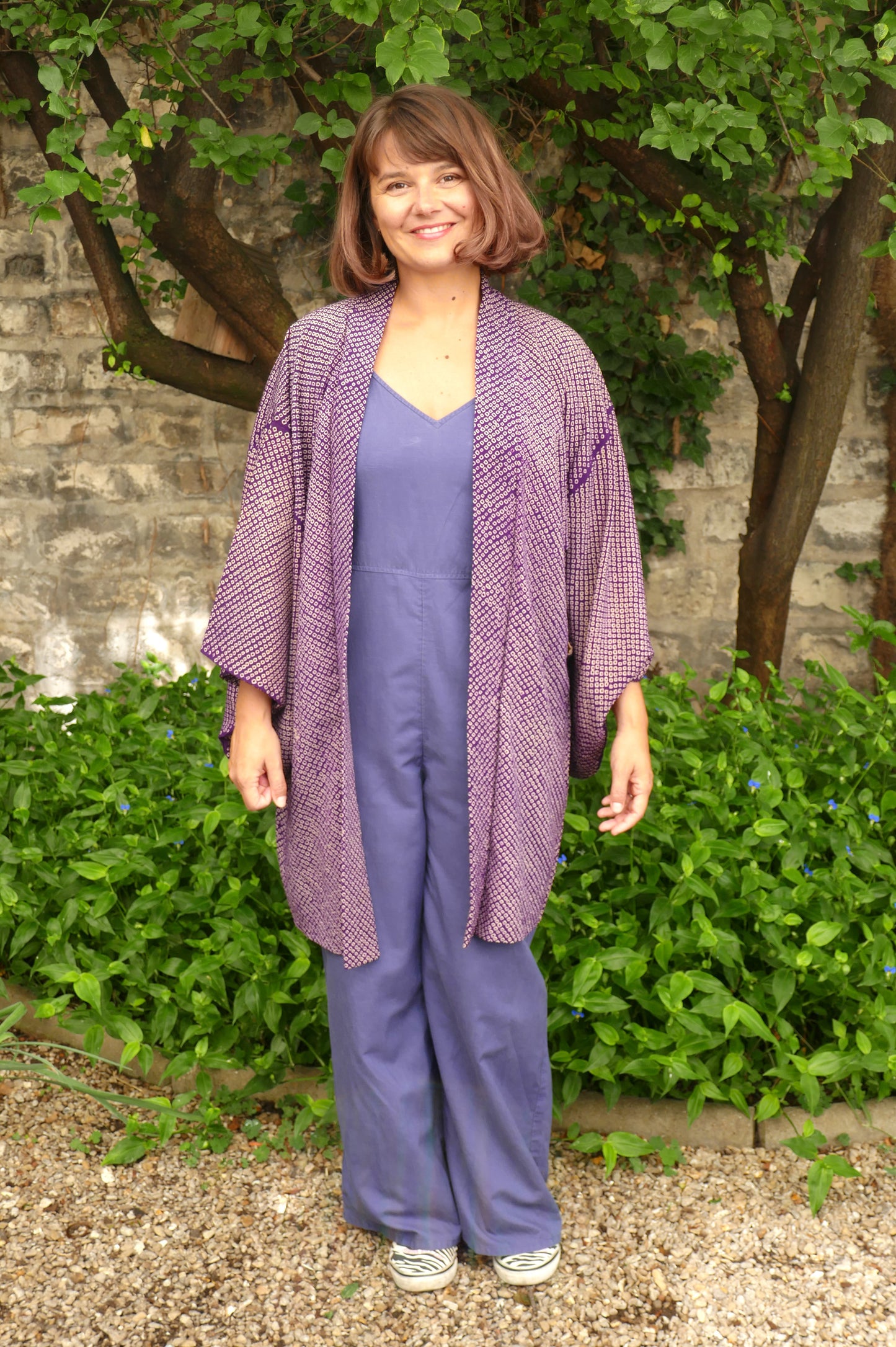 Mikan souriante portant une veste kimono japonaise violet foncé à motifs shibori, ouverte sur une combinaison violette, posant dans un jardin vert. Haori vintage élégant, style japonais authentique.