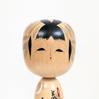 Gros plan sur le visage peint de la kokeshi japonaise Shūhō, coiffure noire et traits traditionnels stylisés.