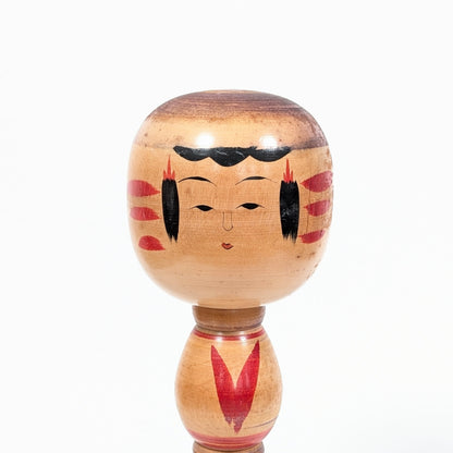 Détail du visage peint d’une kokeshi Ogura Katsushi, avec coiffure noire, ornements rouges et grain naturel du bois