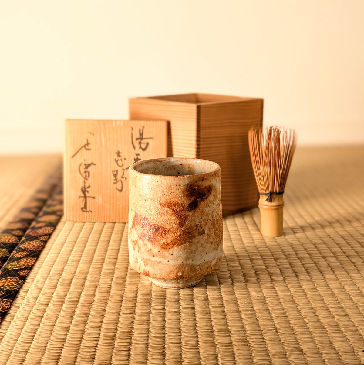 Tasse à thé japonaise Shino sur tatami, accompagnée de sa tomobako en bois calligraphiée et d'un fouet à thé en bambou.