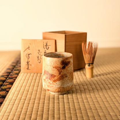 Tasse à thé japonaise Shino sur tatami, accompagnée de sa tomobako en bois calligraphiée et d'un fouet à thé en bambou.